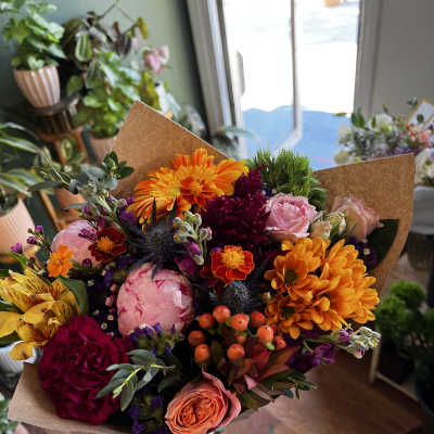 Handheld bouquet of mixed orange, pink, and burgundy flowers in brown paper