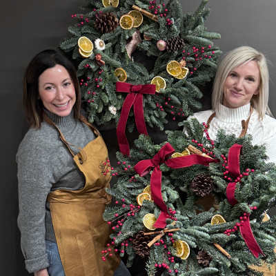 Two women hold decorated evergreen wreaths with red bows and dried citrus slices.
