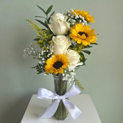 White roses and yellow sunflowers in a clear glass vase with a white ribbon