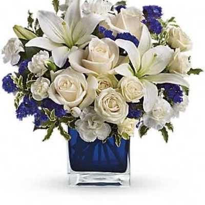 Compact arrangement of white lilies and roses with blue accents in a blue glass cube vase