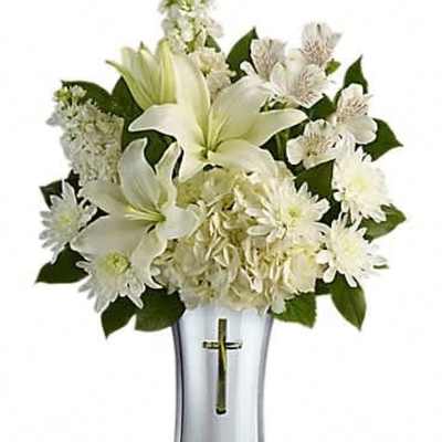 White lilies and mixed white flowers in a tall silver vase with a gold cross emblem