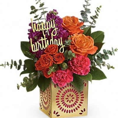 Bright arrangement of orange roses, pink carnations, and purple flowers in a gold cube with a happy birthday sign.