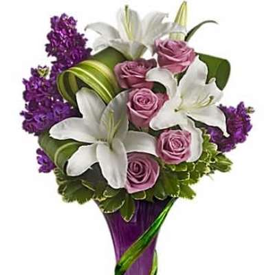 Tall arrangement of white lilies and pink roses with purple blooms in a purple and green glass vase