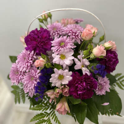 Basket arrangement of pink mums, spray roses, and purple carnations with a white handle.