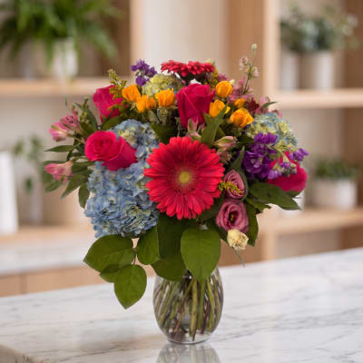 Mixed bouquet in a glass vase with pink roses, blue hydrangeas, and a red gerbera daisy