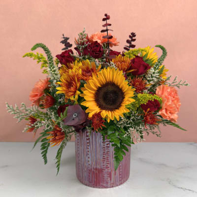 Mixed bouquet with sunflowers, red roses, and peach carnations in a pink vase