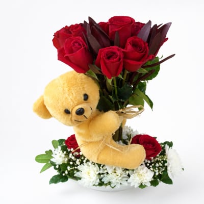 Red roses with a yellow teddy bear and white flowers in a low arrangement