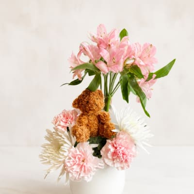 Pink lilies and carnations arranged with a teddy bear in a white vase