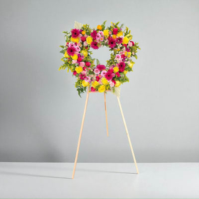 Heart-shaped floral standing wreath on a wooden easel with pink, yellow, and orange blooms
