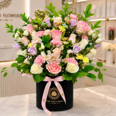 Mixed pink, white, and lavender flowers in a black hatbox with a pink ribbon