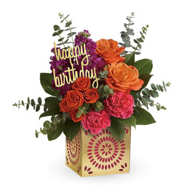 Bright orange roses, pink carnations, and purple blooms in a gold cube vase with a happy birthday pick