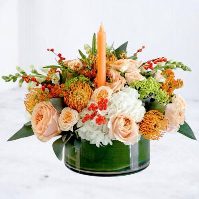 Peach roses and white blooms arranged around an orange candle in a glass vase