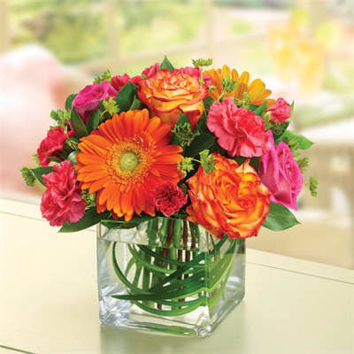 Mixed bouquet of orange, pink, and yellow flowers in a clear square vase