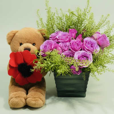 Teddy bear beside a vase of purple roses and greenery