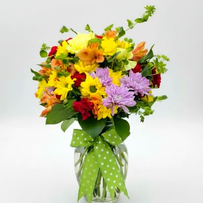 Mixed bouquet of yellow, purple, orange, and red flowers in a glass vase with a green ribbon