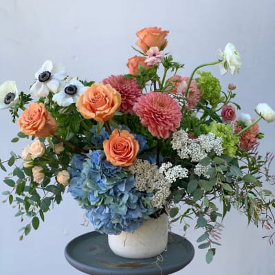 Mixed bouquet with peach roses, pink dahlias, blue hydrangea, and white anemones in a white vase.