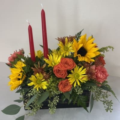 Flower arrangement with sunflowers, orange roses, and red candles in a black container