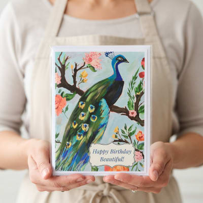 Person holding a birthday card with a painted peacock and flowers
