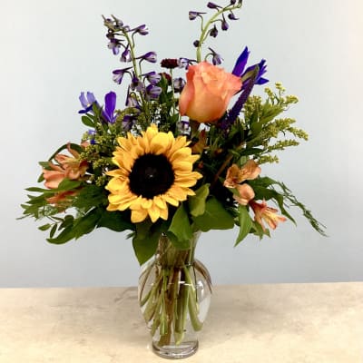 Mixed bouquet with a sunflower, peach rose, and purple flowers in a glass vase