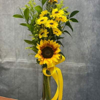 Tall bouquet of yellow sunflowers and small yellow daisies in a clear vase with a yellow ribbon