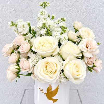 White and blush roses arranged in a white vase with a gold dove emblem