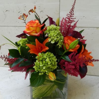 Orange roses and lilies in a clear square vase with green and burgundy accents