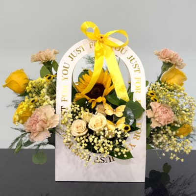 Yellow and peach floral arrangement in a white gift bag with a ribbon handle