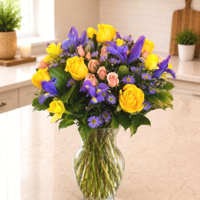 Bouquet of yellow roses, purple irises, and small pink flowers in a clear glass vase