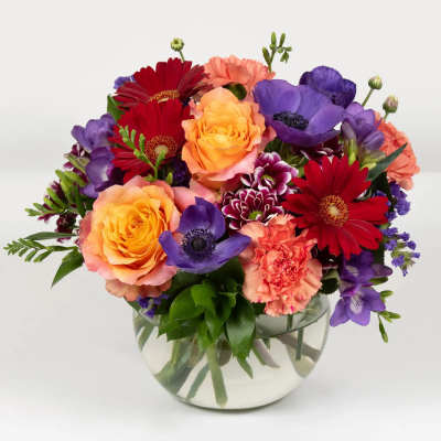 Mixed bouquet of roses, gerbera daisies, carnations, and purple blooms in a glass bowl vase