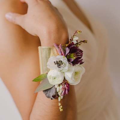 Gold cuff bracelet corsage with white and plum flowers worn on a person's forearm
