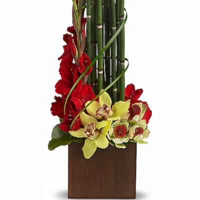 Tall floral arrangement with red flowers, yellow orchids, and bamboo in a wooden vase