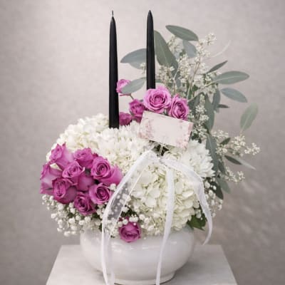 White floral arrangement with purple roses in a white bowl and two black candles