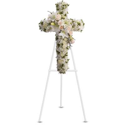 Standing cross-shaped spray of white flowers with lilies and pale peach roses on a white easel stand