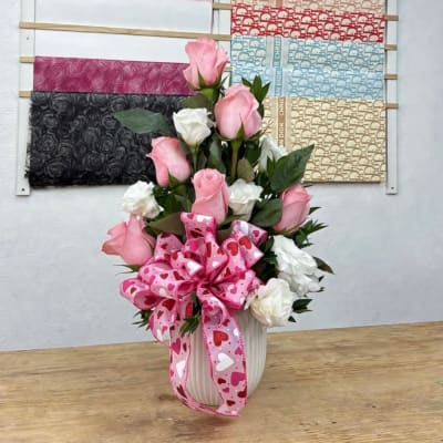 Pink and white roses arranged in a white vase with a heart ribbon