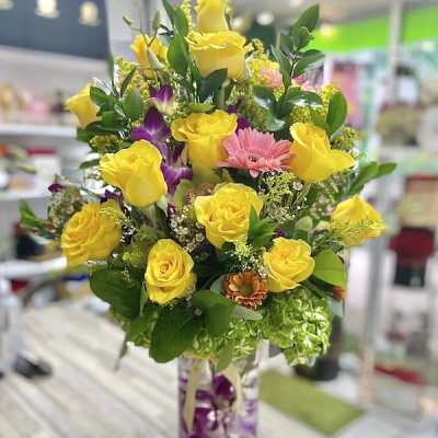 Tall arrangement of yellow roses, pink daisies, and orchids in a clear cylinder vase
