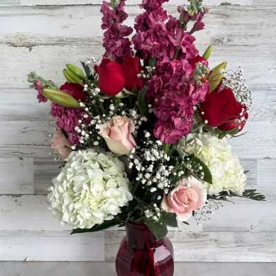Tall arrangement of pink stock, red and pink roses, and white hydrangeas in a red glass vase