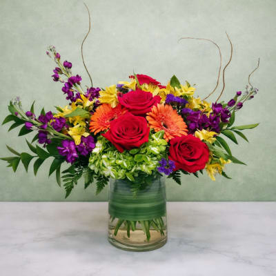 Mixed bouquet of red roses, orange gerberas, and purple flowers in a glass vase