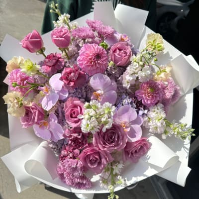 Pink and lavender bouquet with roses, orchids, and mixed blooms wrapped in white paper