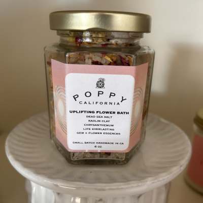 A glass jar of flower bath salts with a gold lid on a white pedestal