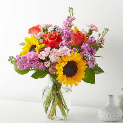 Mixed bouquet with sunflowers, orange roses, and pink blooms in a clear glass vase