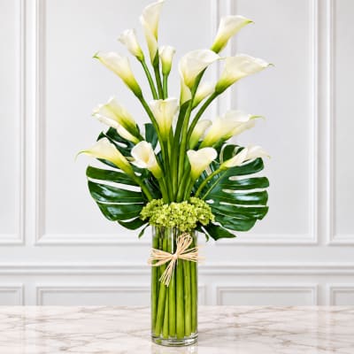 White calla lilies in a tall glass vase with green tropical leaves