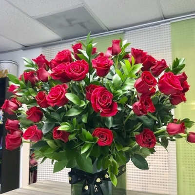 Large arrangement of red roses with abundant greenery in a clear glass cube vase tied with a black ribbon.