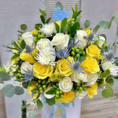 Hatbox arrangement of yellow and white roses with mums, blue thistles, and an It’s a Boy pick.