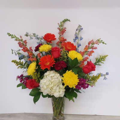 Tall mixed arrangement of red roses, orange gerbera daisies, yellow mums, and white hydrangea in a clear vase