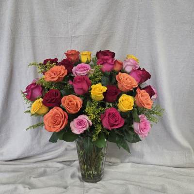 Tall clear vase filled with mixed red, pink, orange, and yellow roses with small yellow filler flowers
