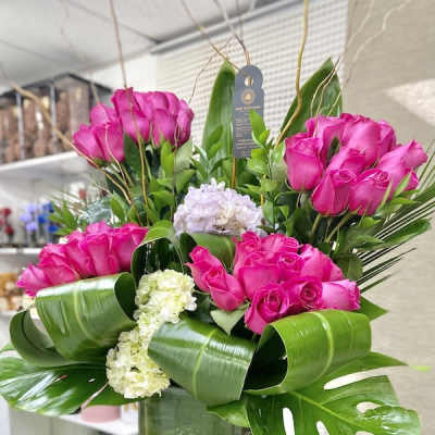 Tall arrangement of hot pink roses with white hydrangeas and large green tropical leaves in a glass cube vase