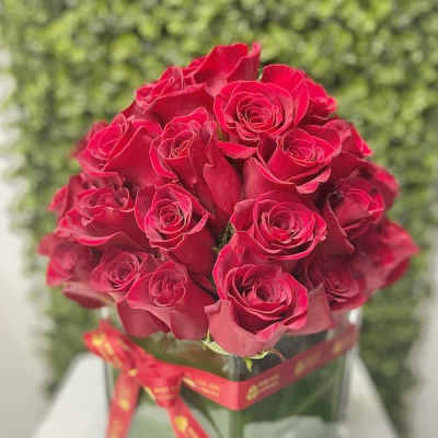 Dense bouquet of red roses arranged in a square glass vase wrapped with a red ribbon.