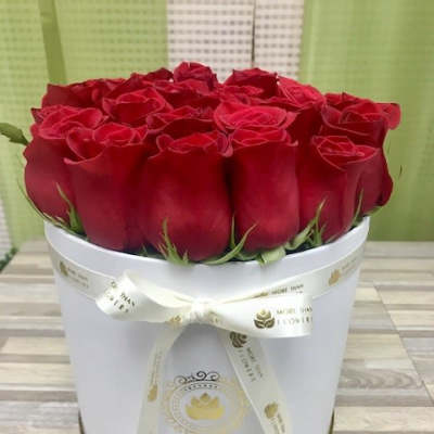 Round white hatbox filled with tightly packed red roses and tied with a cream ribbon bow.