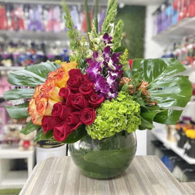 Tall arrangement of red roses, calla lilies, orchids and green hydrangea in a round glass vase with large tropical leaves.