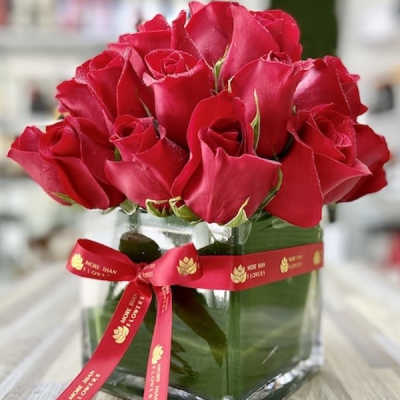 Compact arrangement of red roses in a clear glass cube vase with a red printed ribbon